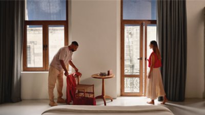 A man adjusting a jacket on a chair and a woman opening a balcony door in a hotel room.