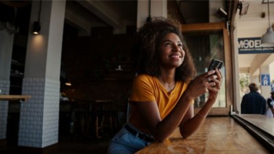 Mulher sorrindo enquanto segura um celular no balcão de uma delicatéssen. 