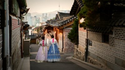 Two women in Seoul, South Korea wearing traditional Hanbok dresses in historic Bukchon Hanok village, with the modern skyline of the city in the distance.