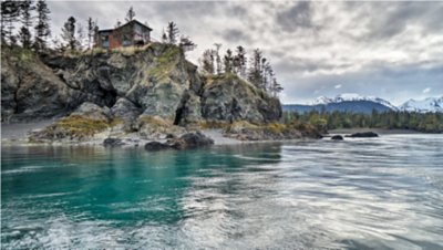 A cabin perched atop a rugged cliff overlooking a turquoise river with distant snow-capped mountains.