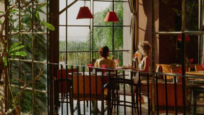 View through windows inside a hotel restaurant, featuring two guests sitting at table and tropical plants and trees outside