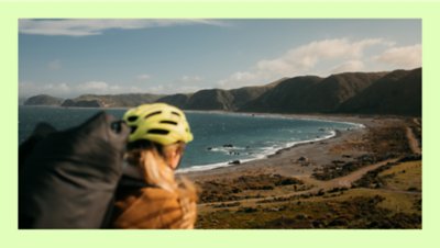 Person wearing a helmet with a backpack overlooking a dramatic coastal landscape with mountains and waves