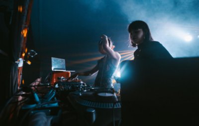Dos DJ actúan en un club nocturno poco iluminado, con humo y puntos de luz que crean una atmósfera misteriosa.