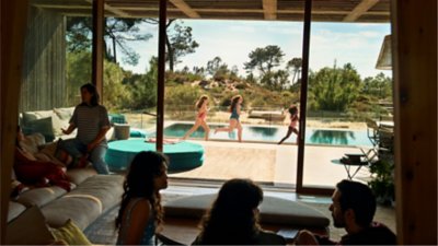 Children in swimsuits run beside an outdoor pool while family members gather and talk inside a modern vacation home with large windows