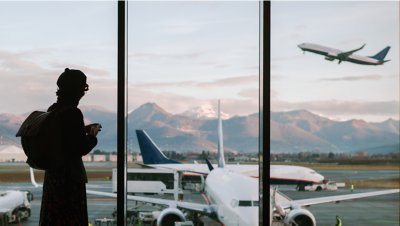 Photograph of a silhouetted female traveler on a mobile device, looking out an airport window with multiple passenger planes in the background.