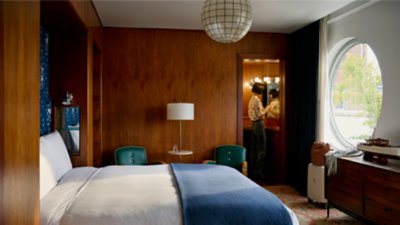 An interior view of a bright hotel room with rich wood paneling. A woman can be seen in the background standing before a small vanity mirror. 