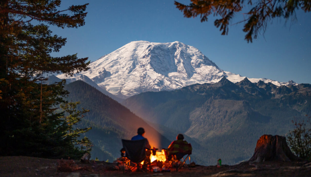 Zwei Menschen sitzen am Lagerfeuer auf einer Lichtung. Im Hintergrund ist ein großer Berg zu sehen.