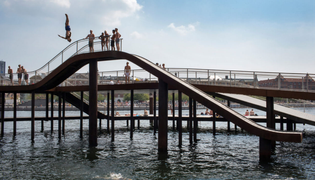 Eine Handvoll Leute stehen auf einer Brücke in der Sonne. Eine Person macht einen Kopfsprung ins Wasser.