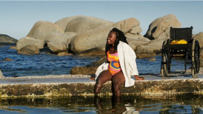  Eine Frau sitzt an einer Uferpromenade in Kapstadt und bereitet sich auf das Schwimmen vor. Ihr Rollstuhl ist im Hintergrund zu sehen.