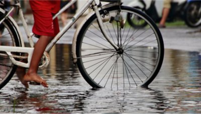  洪水で水浸しになった道路で自転車をこぐ靴を履いていない子供の足のクローズアップ写真。