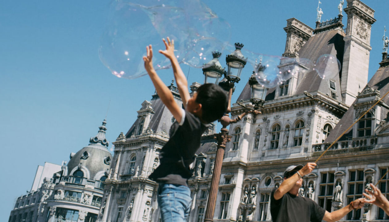 A boy plays with a large bubble in front of some buildings
