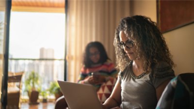 Frau, die mit einem Laptop im Wohnzimmer eines Apartments sitzt, während im Hintergrund eine andere Frau ihr Smartphone nutzt.