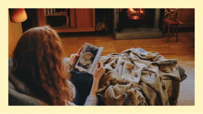 Person with long hair reading and relaxing on a sofa covered with a blanket in front of a fireplace
