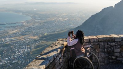 Une femme en fauteuil roulant prend une photo de la vue depuis une montagne qui surplombe la ville en contrebas et le bord de l’océan.