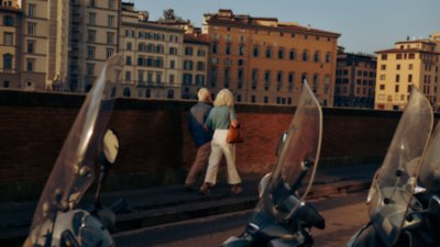 A view of an older couple from behind, touring the city with scooters in the foreground and buildings in the background.