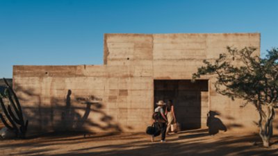 Duas pessoas carregando bagagens enquanto caminham para a entrada com fachada em concreto do hotel Paradero Todos Santos no México.