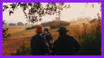 Photograph of two men with a large camera, filming a small cabin and truck in a meadow..