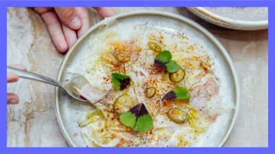 Overhead photograph of a plate of fish carpaccio and hands holding a spoon.