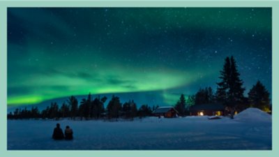  Foto von zwei Personen, die im Schnee sitzen und die Nordlichter betrachten, mit einer gemütlichen Blockhütte im Hintergrund.