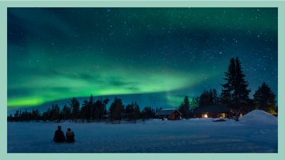  Photograph of two people sitting in snow looking at the northern lights with a cozy log cabin in the background.