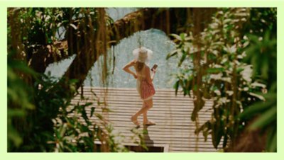  Photograph taken through the limbs of a tree of a woman in a sunhat walking near a body of water, holding her smart phone.