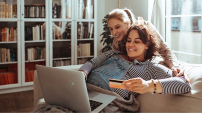 Two women in a living room making payment on a laptop; one who is pregnant, sits on the couch with the laptop on her legs, holding a credit card in one hand, while the other woman leans on her shoulder.